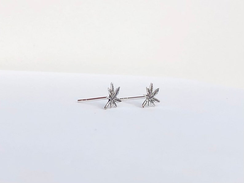 May include: A pair of silver-colored stud earrings. Each earring features a detailed cannabis leaf design. The earrings are displayed on a white surface, with a simple and clean aesthetic. The earrings are small and delicate.