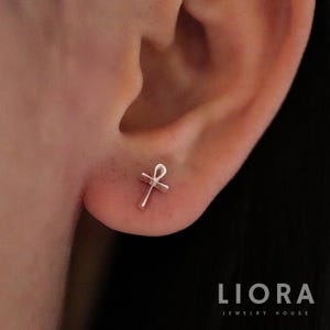 May include: A small, silver ankh stud earring. The earring is a minimalist design, featuring the ankh symbol, a cross with a loop at the top. The earring is shown on an ear, with the Liora Jewelry House logo in the bottom right corner.