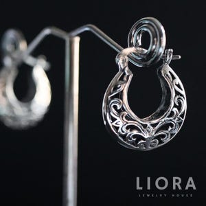 May include: Silver hoop earrings featuring a delicate, intricate floral design. The earrings are displayed on a stand against a black background.