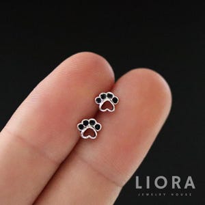 May include: Two small silver paw print stud earrings with black accents. The earrings are displayed on a finger against a dark background. The earrings are from the Liora Jewelry House.