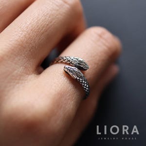 Puede incluir: Un anillo de serpiente plateado con escamas detalladas envuelve un dedo. La cabeza y la cola de la serpiente se encuentran, creando un diseño único y elegante. El anillo es una joya de LIORA JEWELRY HOUSE.