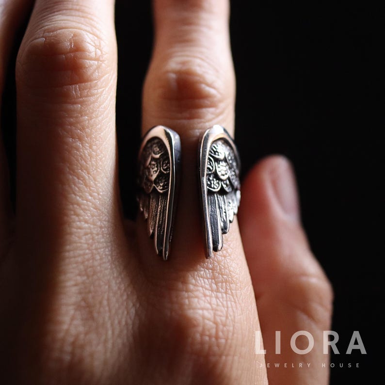 May include: A silver-colored ring with an open design, featuring detailed wing motifs on each side. The wings are intricately carved with feather and texture details. The ring is worn on a finger, showcasing the jewelry's design.