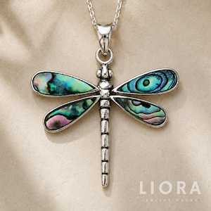 May include: A silver-toned dragonfly pendant necklace featuring iridescent abalone shell inlays within the four wings. The dragonfly body has a textured, segmented appearance, and the piece hangs from a delicate silver-toned chain.