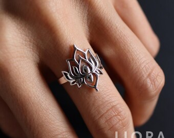 Puede incluir: Un anillo de plata con forma de flor de loto. El anillo presenta un diseño calado de una flor de loto con pétalos detallados. La joya es de Liora Jewelry House.