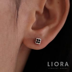 May include: A close-up of a person's ear wearing a silver earring with a black square design. The earring is a small, geometric shape with a black center and a silver border.