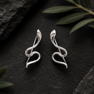 May include: A pair of silver-toned ear climbers shaped like stylized snakes. Each earring features a smooth, polished metallic finish with a curved, serpentine body that follows the contour of the earlobe, ending in a pointed tail and head.