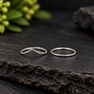 Pode incluir: Dois anéis de tom prateado com uma superfície texturizada e com contas. Um anel apresenta um design em forma de V, enquanto o outro é uma banda circular fina e simples. Ambos os anéis têm um acabamento oxidado escuro.