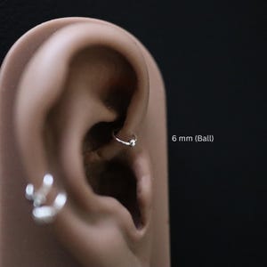 May include: A close-up of a person's ear with a small silver hoop earring with a 6 mm ball. The earring is in the upper cartilage of the ear.