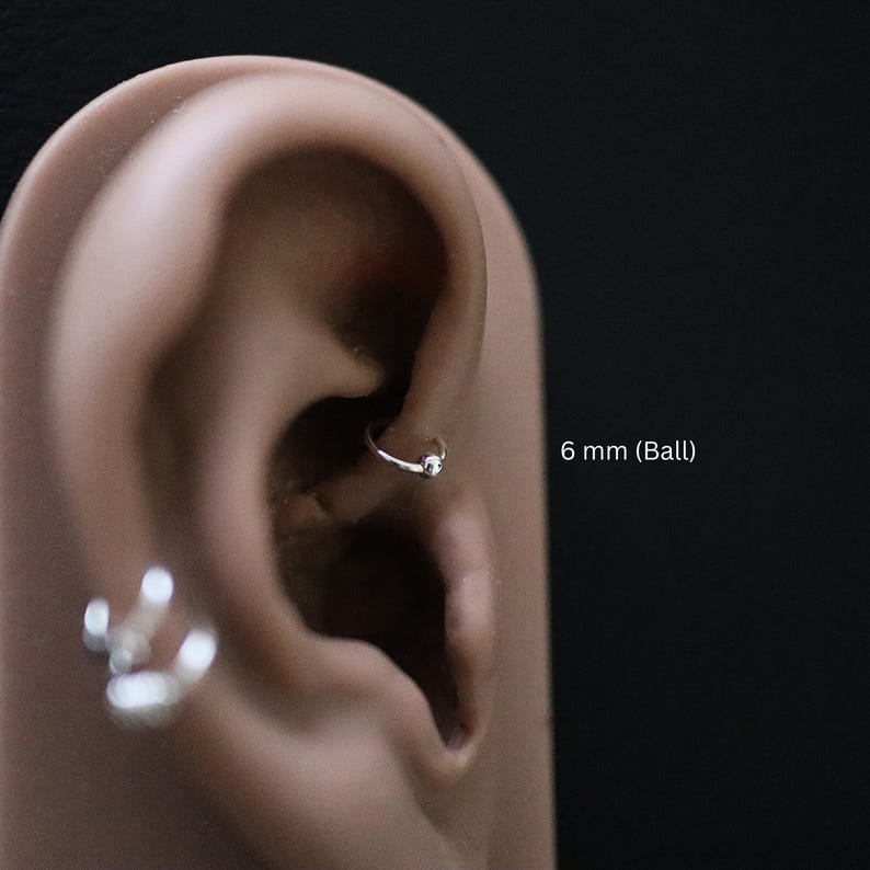 May include: A close-up of a person's ear with a small silver hoop earring with a 6 mm ball. The earring is in the upper cartilage of the ear.