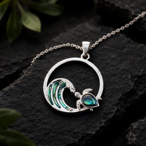 Può includere: Una collana con ciondolo in argento caratterizzata da una cornice circolare contenente un'onda oceanica stilizzata e una tartaruga marina. L'onda e il guscio della tartaruga sono intarsiati con conchiglia di abalone iridescente nei toni del verde, blu e viola.