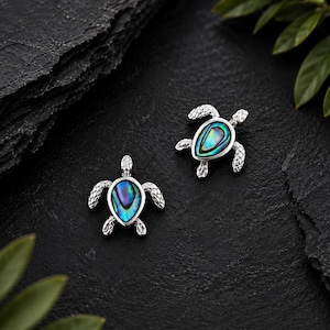 Op de afbeelding: Twee zilverkleurige oorbellen in de vorm van een schildpad met inlays van iriserende abalone-schelp op de schilden. De metalen oppervlakken hebben een gestructureerd, schubachtig patroon op de flippers en de kop van de schildpad.