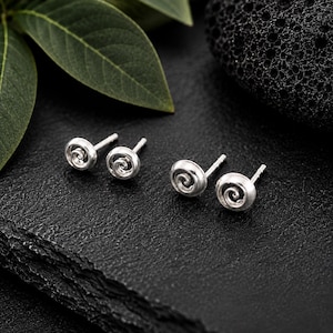 Op de afbeelding: Twee paar zilveren oorknopjes, elk met een cirkelvormig ontwerp met een centraal spiraalpatroon. De oorbellen hebben een gepolijste metalen afwerking en zijn voorzien van standaard stekers voor bevestiging aan de oren.