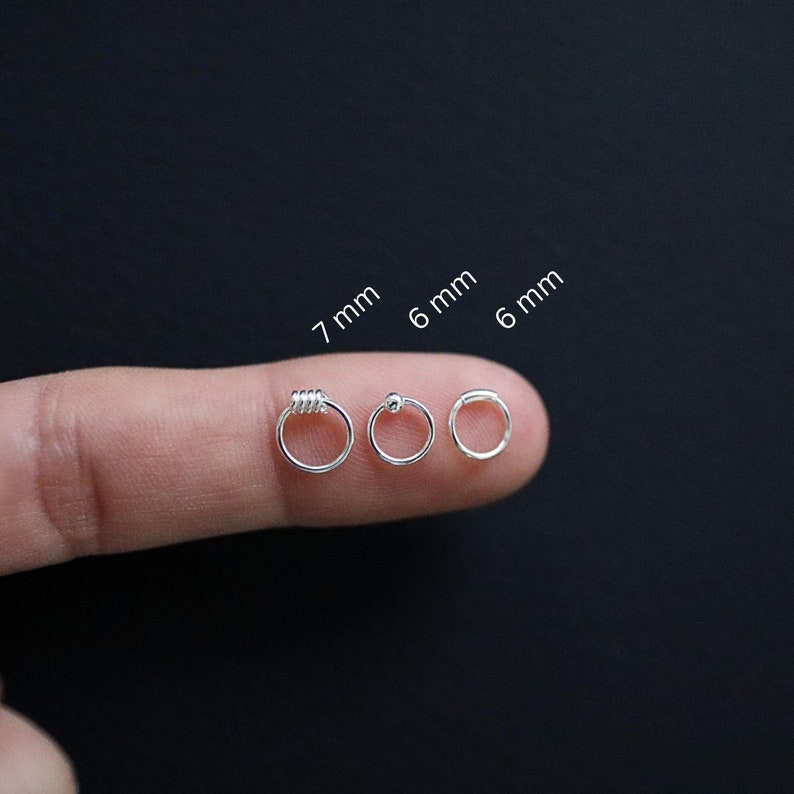 May include: Three silver nose rings in different sizes: 7 mm, 6 mm, and 6 mm. The rings are shown on a finger, with the largest ring on the left and the smallest ring on the right.