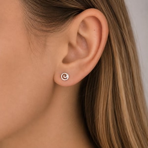 Op de afbeelding: Een kleine, zilverkleurige oorsteker met een eenvoudig, cirkelvormig spiraalontwerp. De oorsteker zit in de oorlel en heeft een gepolijste metalen afwerking en een opgerolde vorm die zorgt voor een strak en ingetogen uiterlijk.