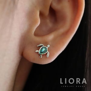 Puede incluir: Un pendiente de botón de tortuga plateada con incrustaciones de abulón turquesa y verde. El pendiente presenta aletas y una cabeza texturizadas. El pendiente se muestra en una oreja.