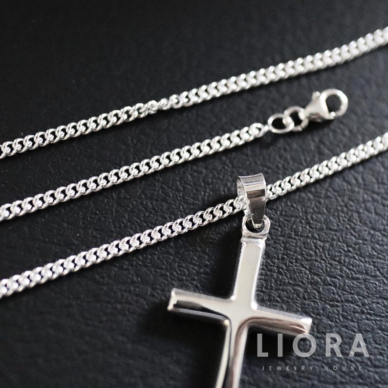 May include: A silver chain necklace with a silver cross pendant. The chain is a curb chain style. The cross pendant is simple and has a smooth finish. The necklace is displayed on a black surface. Liora Jewelry House is printed on the surface.
