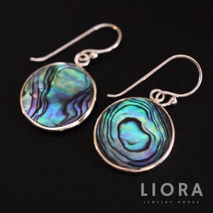 Peut inclure: Une paire de boucles d'oreilles rondes en coquillage d'ormeau avec des crochets d'oreilles en argent sterling. Les coquillages irisés présentent des nuances de bleu, de vert et de violet. Les boucles d'oreilles sont posées sur un fond noir.