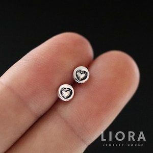 Puede incluir: Un par de pequeños pendientes de botón plateados. Cada pendiente presenta un diseño de corazón dentro de un marco circular. Los pendientes se muestran en un dedo sobre un fondo oscuro. La marca "LIORA JEWELRY HOUSE" es visible.