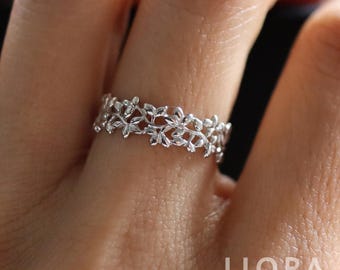 May include: A silver ring featuring a delicate floral design. The ring is crafted with intricate details, showcasing a repeating pattern of leaves and small flowers. The jewelry is a close-up shot, highlighting the ring's texture and design.