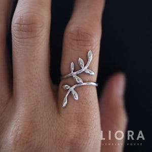 Puede incluir: Un anillo plateado con un delicado diseño de hojas. El anillo se envuelve alrededor del dedo con dos bandas, y las hojas están agrupadas en la parte superior. La joya es de Liora Jewelry House.