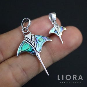 Puede incluir: Dos colgantes plateados de raya con alas de concha de abulón iridiscente. Los colgantes tienen un diseño detallado y un lazo para una cadena. El fondo es oscuro, resaltando las joyas.