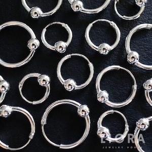 May include: A collection of silver hoop earrings with a small, round bead accent. The earrings are arranged on a dark surface, showcasing their reflective, polished finish. The jewelry is from the Lora Jewelry House.