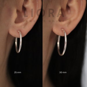 Puede incluir: Dos pendientes de aro de plata, uno de 25 mm y el otro de 30 mm, se muestran en los l&oacute;bulos de las orejas de una persona. Los pendientes son simples y elegantes.