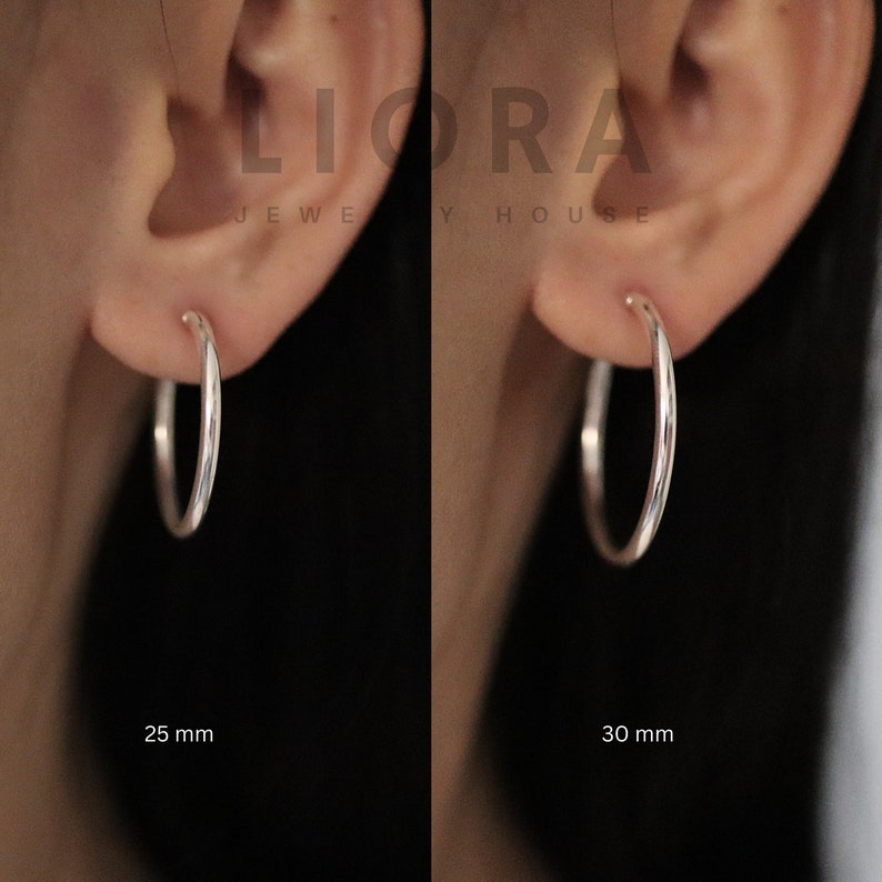 Puede incluir: Dos pendientes de aro de plata, uno de 25 mm y el otro de 30 mm, se muestran en los l&oacute;bulos de las orejas de una persona. Los pendientes son simples y elegantes.
