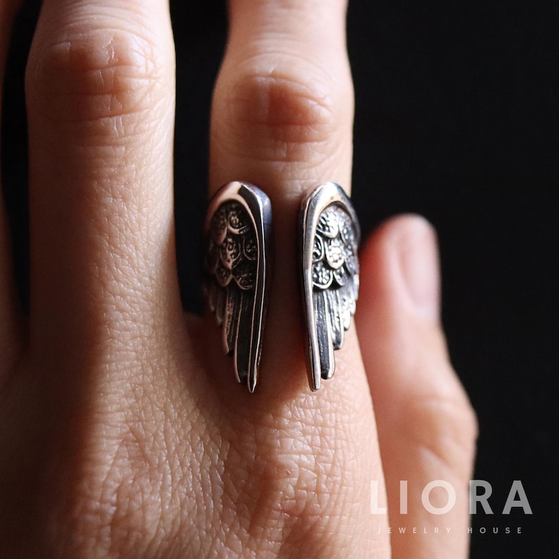 May include: A silver-colored ring with an open design, featuring detailed wing motifs on each side. The ring is worn on a finger, showcasing the intricate feather detailing. The jewelry is from LIORA JEWELRY HOUSE.