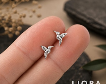 Könnte beinhalten: Ein Paar kleine Kolibri-Ohrstecker in Silberoptik. Jeder Ohrstecker zeigt einen detaillierten, polierten Metallvogel im Flug mit ausgebreiteten Flügeln und einem spitzen Schnabel. Das Design ist kompakt und weist ein metallisches Finish auf.