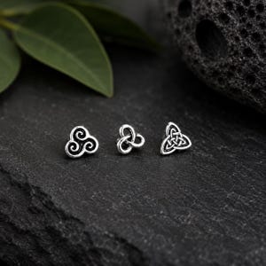 Op de afbeelding: Drie zilverkleurige oorknopjes met verschillende ontwerpen: een triskelion-spiraal, een kleine triquetra-knoop en een grotere driehoekige Keltische knoop. Elk sieraad heeft een geoxideerde afwerking die de gedetailleerde metalen vormen benadrukt.