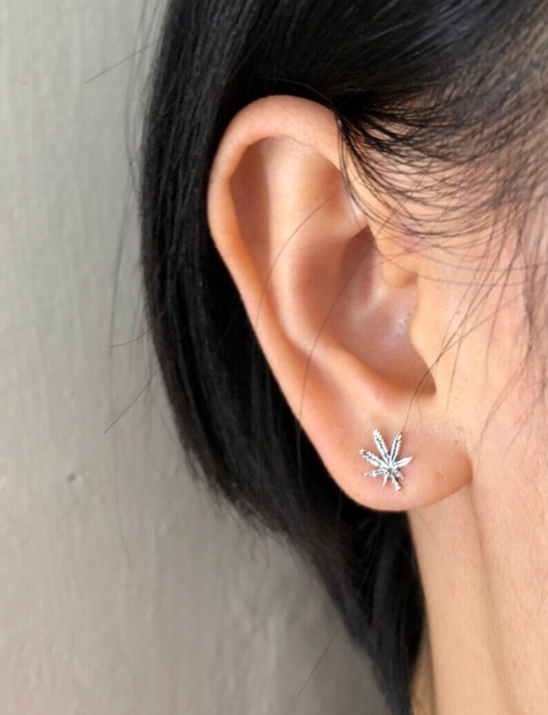 May include: A silver-colored stud earring in the shape of a cannabis leaf is shown on an ear. The earring has detailed veining and a shiny finish. The ear is in focus against a blurred background.