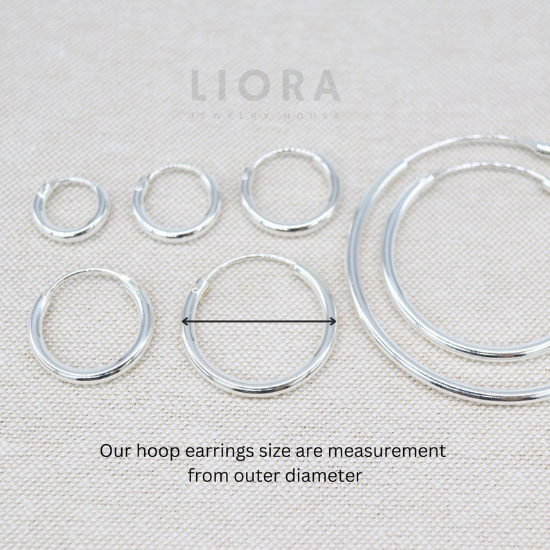Puede incluir: Cinco pendientes de aro de plata de diferentes tama&ntilde;os. Los pendientes est&aacute;n dispuestos en una fila del m&aacute;s peque&ntilde;o al m&aacute;s grande. El pendiente m&aacute;s grande tiene un di&aacute;metro de aproximadamente 5 cm. El texto "LIORA JEWELRY HOUSE" es visible en la parte superior de la imagen.