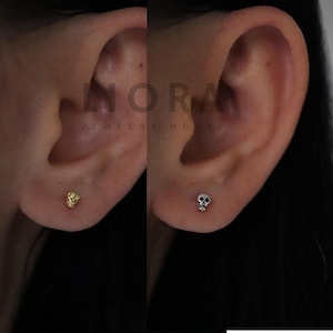 May include: Close-up of a person's ear with two small skull earrings. The earring on the left is gold and the earring on the right is silver.