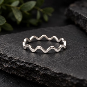 Puede incluir: Un anillo fino de color plateado que presenta un patrón ondulado o en zigzag continuo y repetitivo a lo largo de toda la circunferencia de la banda. El metal tiene un acabado liso y pulido que refleja la luz, creando una forma circular.