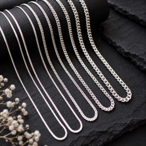 May include: A collection of six silver-toned metal chains arranged in a row, each featuring a distinct link thickness. The chains are displayed in a gradient of sizes, ranging from a thin, delicate style to a thicker, more robust curb link design.
