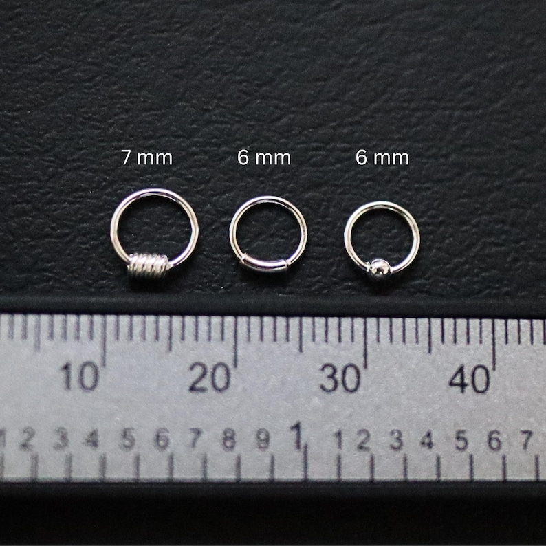 May include: Three silver hoop earrings of different sizes, measured in millimeters, displayed on a ruler. The earrings are 7 mm, 6 mm, and 6 mm in diameter.