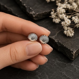 May include: A pair of small, silver-toned stud earrings shaped like miniature seashells. Each earring features a fan-like design with delicate, radiating ridges that mimic the natural texture of a scallop shell, finished with a polished metallic sheen.
