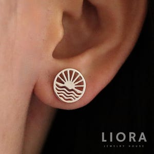 May include: A silver stud earring with a circular design featuring a sun with radiating lines above wavy lines. The earring is worn on an ear. The jewelry is from Liora Jewelry House.