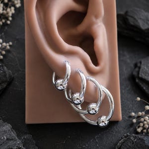 Peut inclure: Un ensemble de quatre boucles d'oreilles créoles argentées, chacune ornée d'une perle sphérique polie. Les anneaux sont de tailles graduées et disposés en rangée, présentant une finition métallique lisse et réfléchissante sur les cercles.