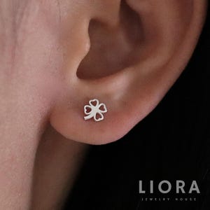 May include: A close-up of a person's ear with a small silver four-leaf clover earring. The earring is a delicate, open design with a heart shape for each leaf.
