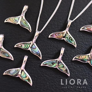 Op de afbeelding: Een verzameling zilveren walstaart hangers met iriserende abalone schelp inlegwerk. De hangers worden getoond op een zwarte ondergrond met een zilveren kettingketting. De afbeelding bevat de tekst "LIORA JEWELRY HOUSE".