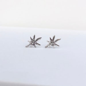 May include: A pair of silver-colored stud earrings, each shaped like a cannabis leaf. The earrings are detailed with veining and are set against a white background. The earrings are small and delicate.