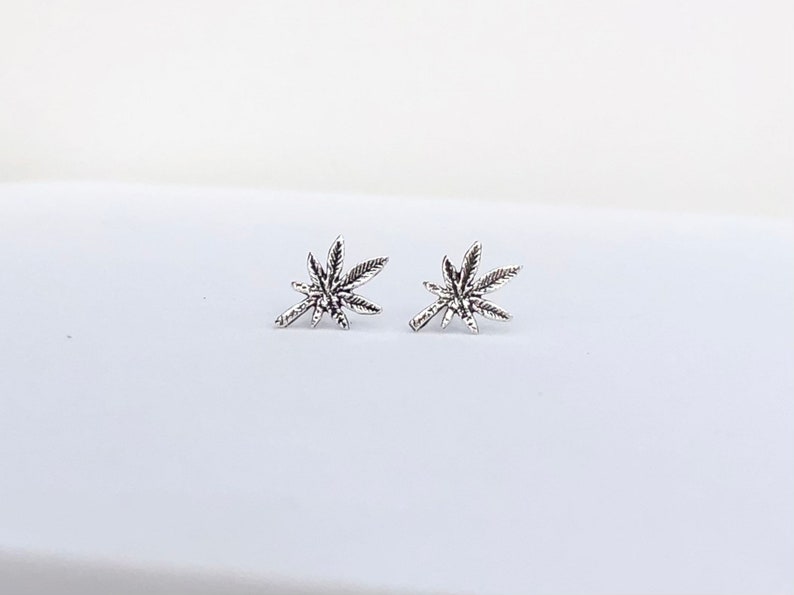 May include: A pair of silver-colored stud earrings, each shaped like a cannabis leaf. The earrings are detailed with veining and are set against a white background. The earrings are small and delicate.