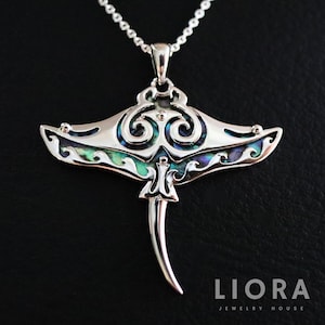 May include: A silver pendant necklace with a manta ray design. The pendant has swirling patterns and iridescent abalone shell inlays. The necklace is on a delicate chain. The brand name "LIORA" is visible at the bottom.