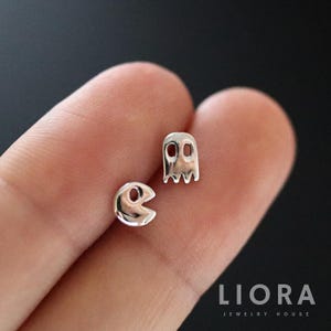 Könnte beinhalten: Ein Paar silberfarbene Ohrstecker mit Pac-Man und Geister-Motiv. Die Ohrringe sind klein und glänzend, mit einem minimalistischen Design. Der Hintergrund ist dunkel und verschwommen. Schmuck im Retro-Stil.