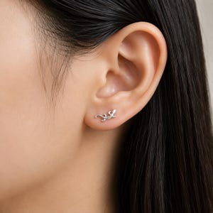 Peut inclure: Une petite boucle d'oreille à clou argentée en forme de lézard, présentant une surface texturée et une queue incurvée, placée sur le lobe de l'oreille. La boucle d'oreille a une finition métallique et une silhouette détaillée imitant un reptile.