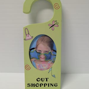 May include: A light green wooden door hanger with a circular opening for a 10cm x 7.5cm photo. The hanger has a curved top and is decorated with pink and black illustrations of a purse, dragonfly, and high heel. The words "OUT SHOPPING" are printed on the bottom.