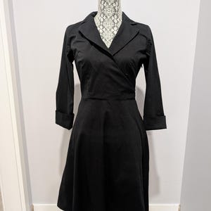 May include: A black wrap dress with a collared neckline and three-quarter sleeves. The dress has a fitted bodice and a flared skirt, displayed on a mannequin. The dress is made of a solid black fabric.