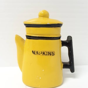 Peut inclure: Porte-serviettes en céramique jaune en forme de cafetière avec une poignée noire et une bordure noire. Le porte-serviettes porte l'inscription "NAPKINS" sur le devant.